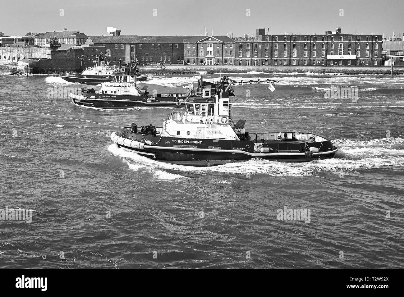 Black And White Photo Of 3 SERCO LTD, Tugs Assisting The Royal Navy Aircraft Carrier, HMS QUEEN ELIZABETH As She Departs Portsmouth Harbour, UK Stock Photo