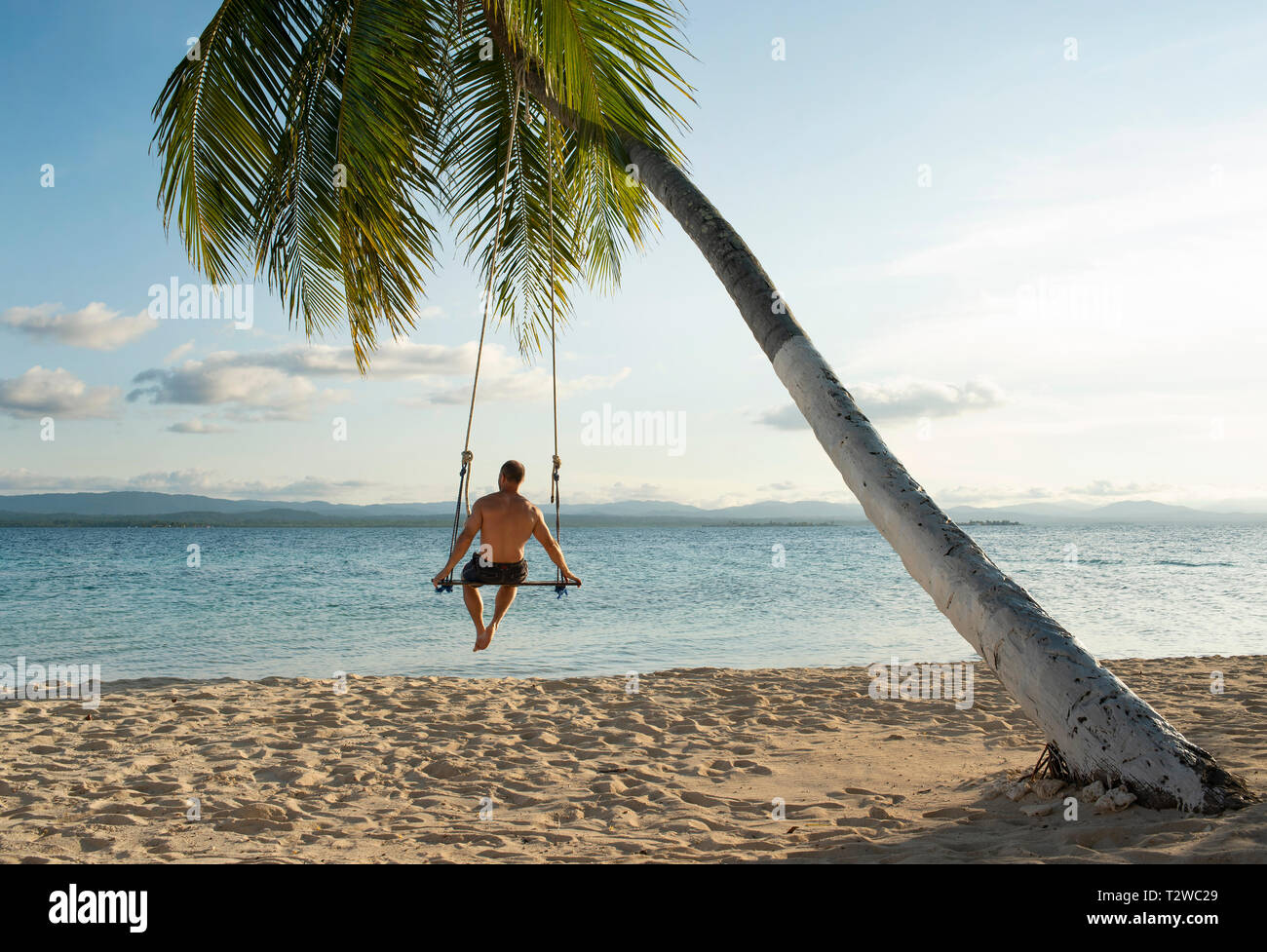 Rear view of man in a beach swing attached to a palm tree in San Blas Islands. Travel destination, lifestyle / holiday concept. Panama, Oct 2018 Stock Photo