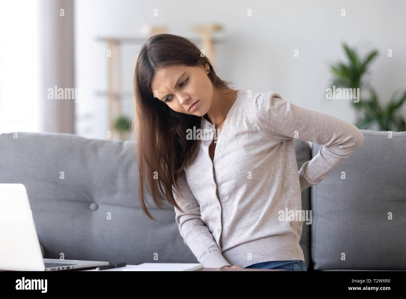 Unhealthy young woman sitting on couch suffers from back pain Stock
