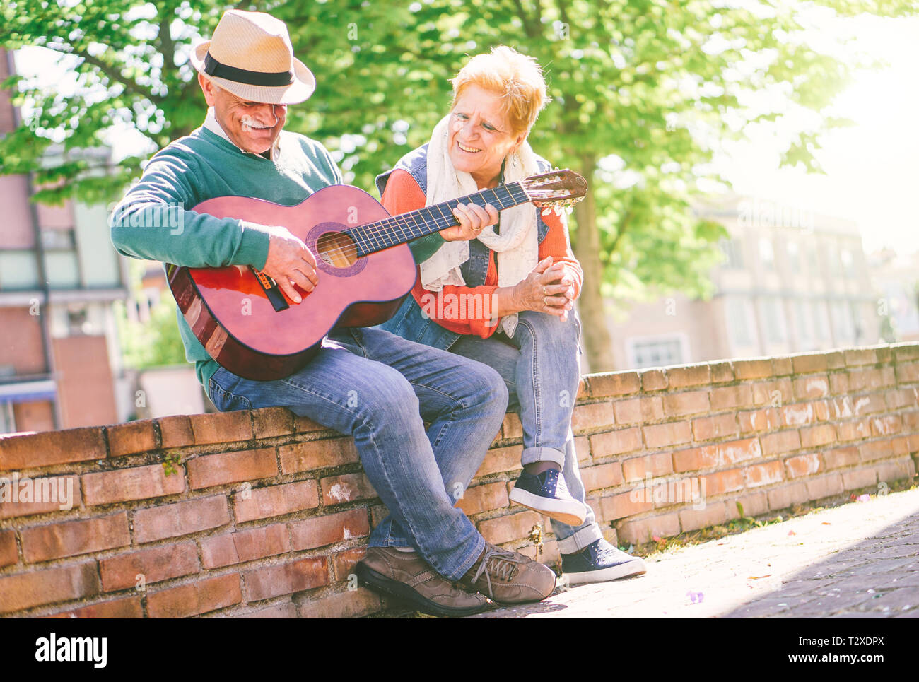 Happy senior couple playing a guitar while sitting outside on a wall on a sunny day - Concept of active elderly having fun with guitar Stock Photo