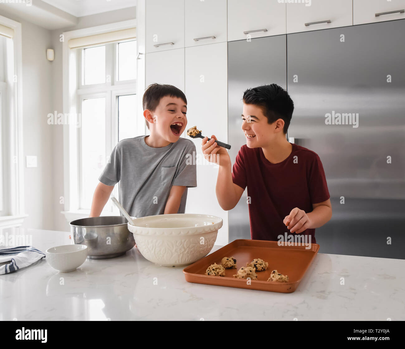 Older boy feeding cookie dough to young boy in modern kitchen Stock Photo