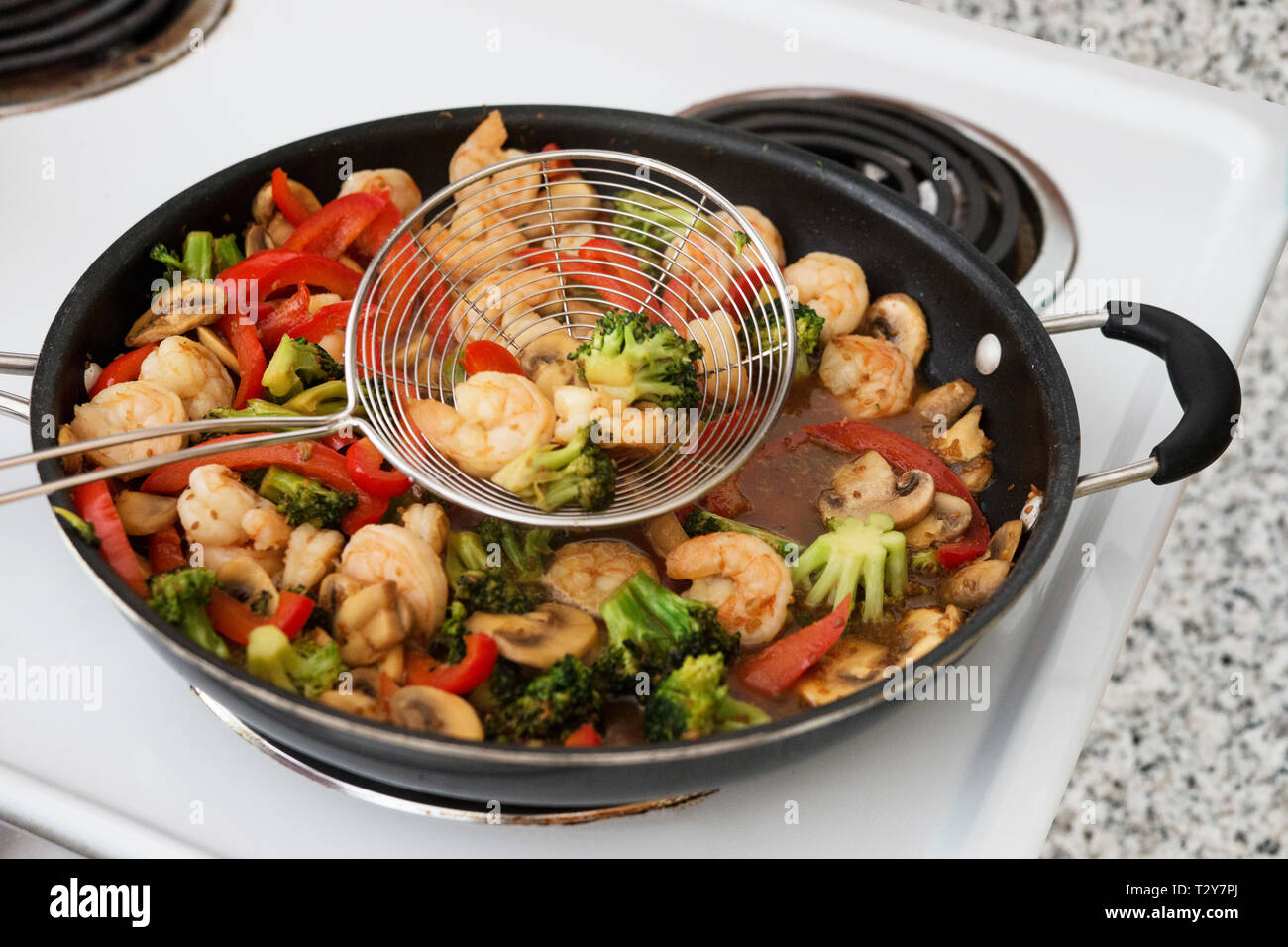 A stir fry of shrimp, mushrooms, broccoli, and red peppers in a skillet on an electric stove with a metal skimmer spoon. Stock Photo