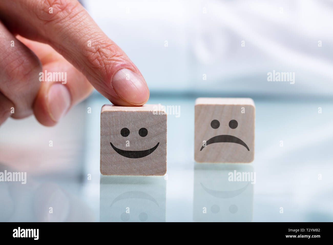 Close-up Of Person's Hand Holding Wooden Block With Happy And Unhappy Face On Reflective Desk Stock Photo