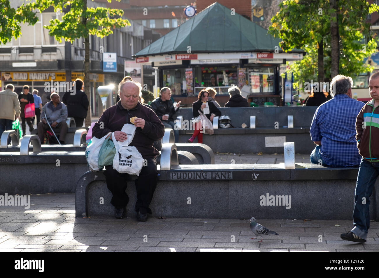 Older man sitting on a concrete bench eating a pie in an urban pedestrican precinct Stock Photo