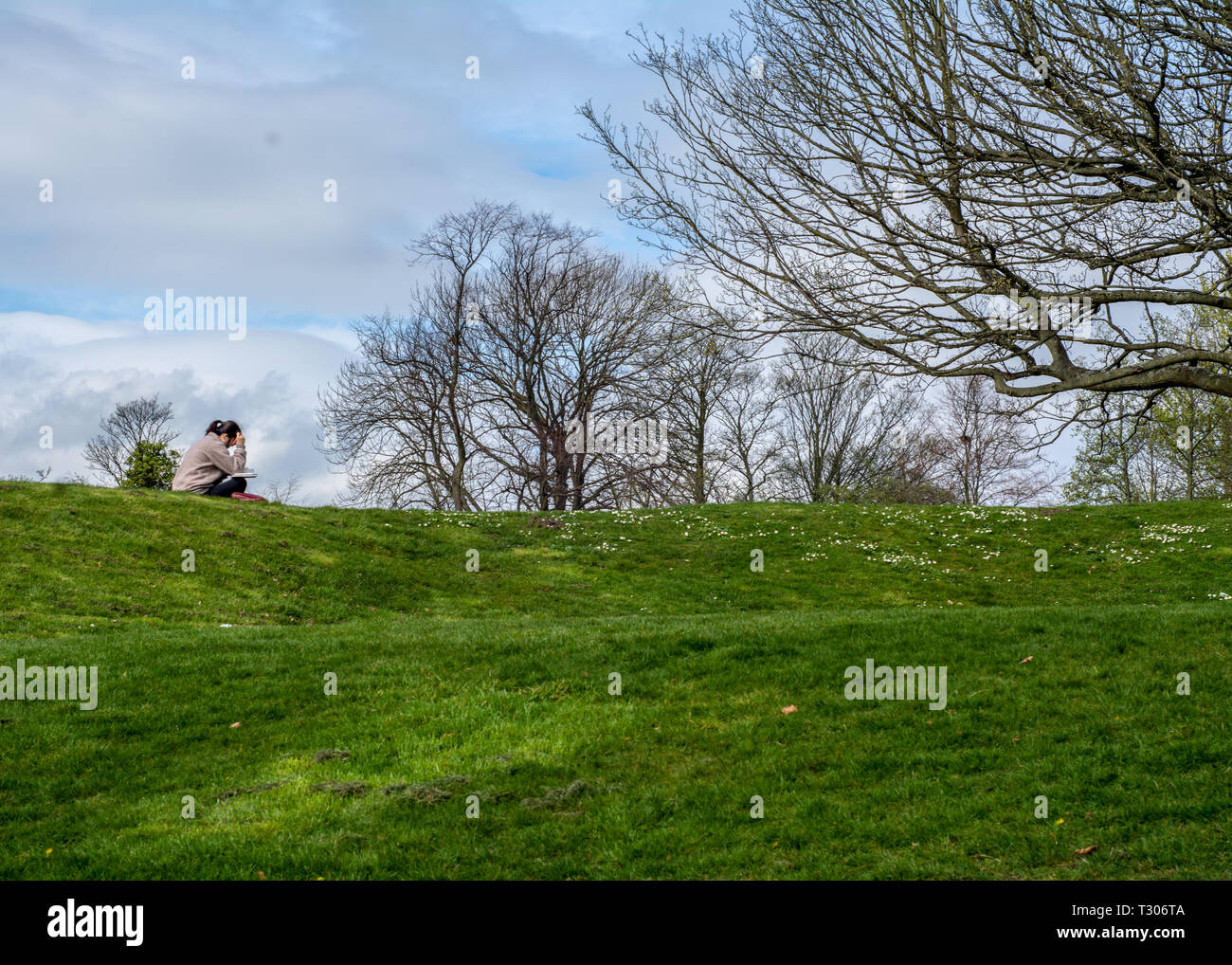 Huddersfield,UK, April 5 2018. A woman sits reading on the grass on a warm spring day at Greenhead Park. Stock Photo