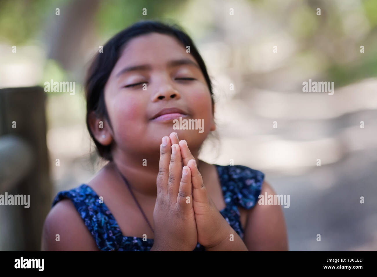 A native young girl with hands together in prayer, in an outdoor setting praying to God with a subtle smile. Stock Photo