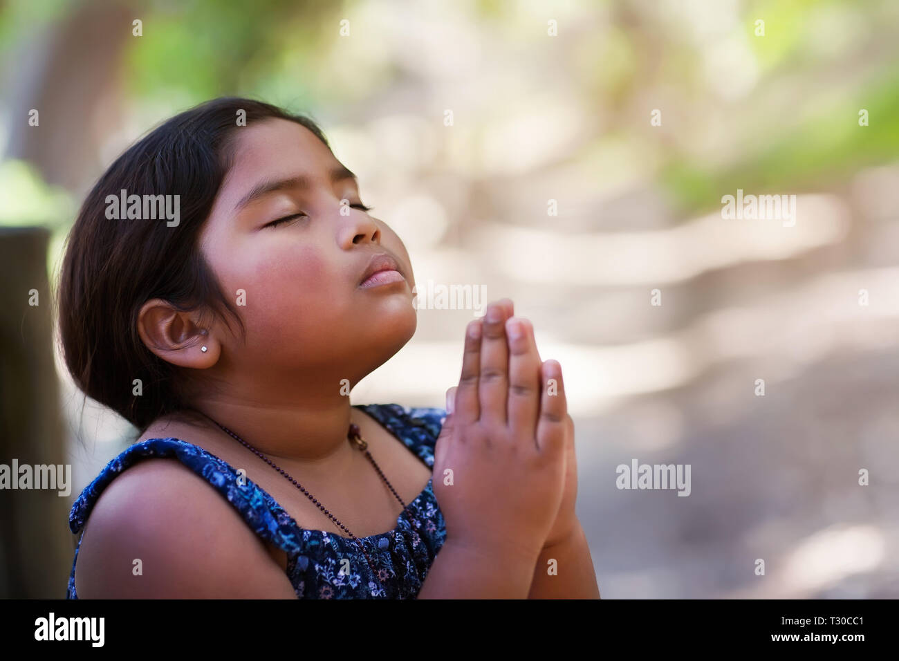 A young girl praying with hands together in reverence to God, wearing conservative clothing and in an outdoor setting. Stock Photo