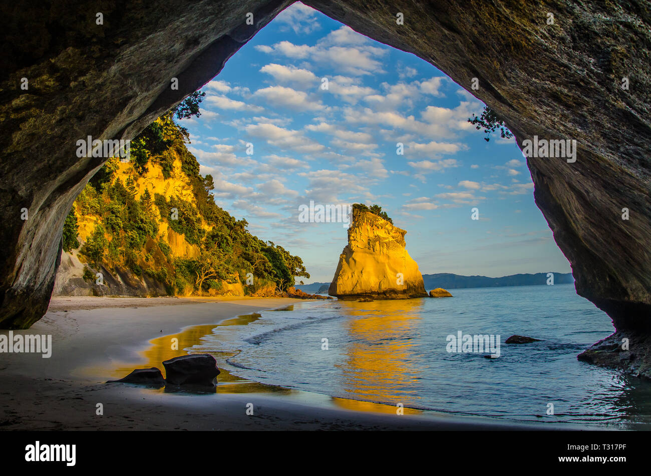 Sunrise at Cathedral Cove with blue sky with white clouds above and reflection in the sea, golden hour. Stock Photo