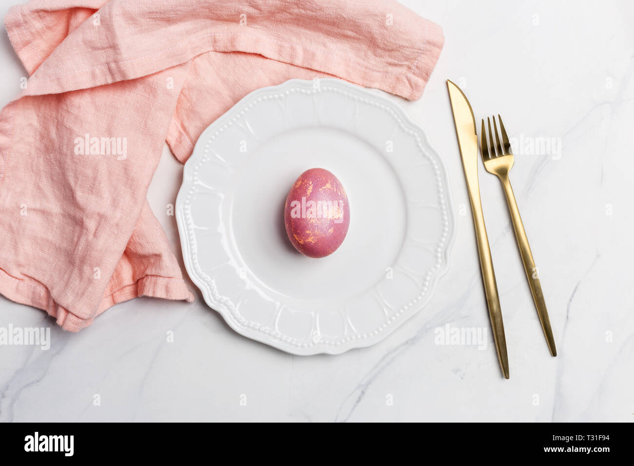 Easter table, place setting. Easter egg, pink color painted in a white plate, pink napkin, golden tableware on white marble background, horizontal, co Stock Photo