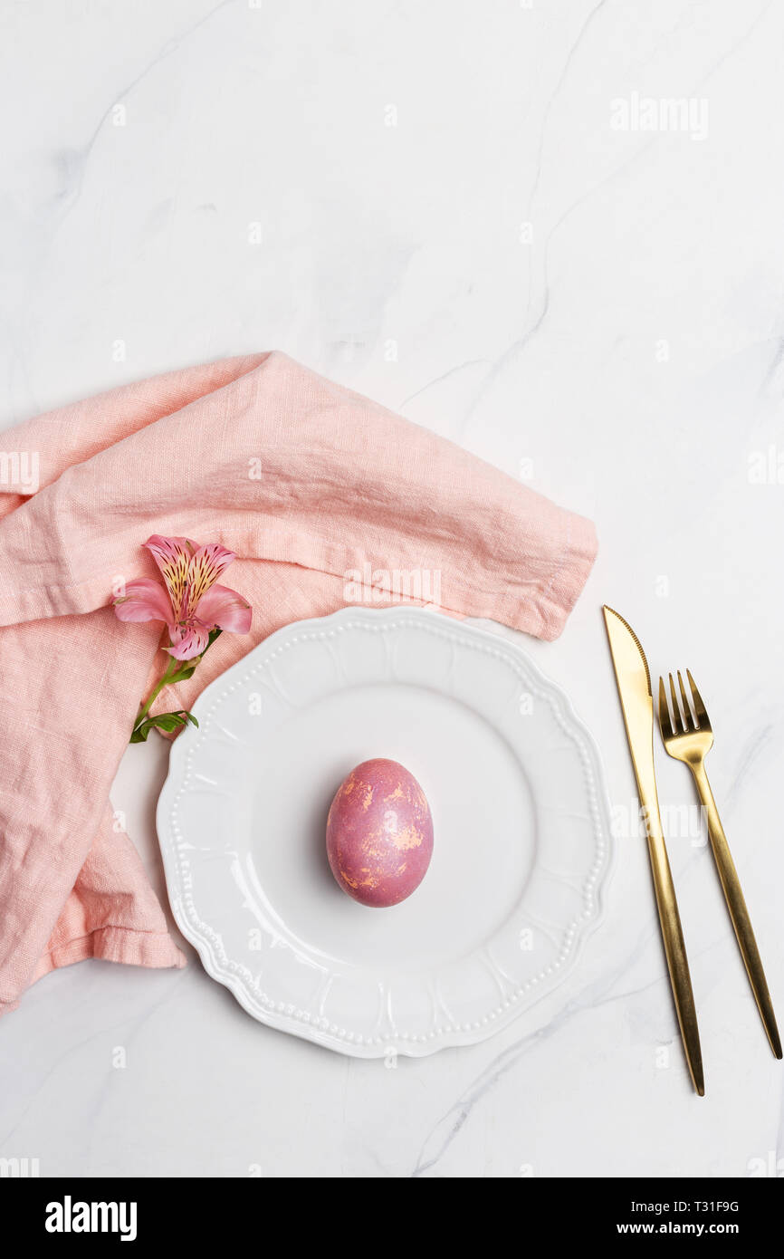Easter table, place setting. Easter egg, pink color painted in a white plate, pink napkin, golden tableware on white marble background, vertical, copy Stock Photo