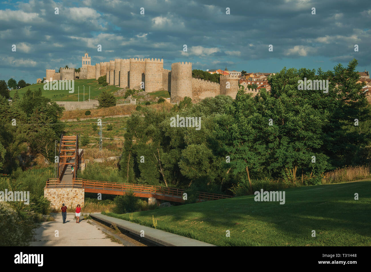 People on wooden bridge over the Adaja River and wall around Avila on top of hill. With an imposing wall around the gothic city center in Spain. Stock Photo