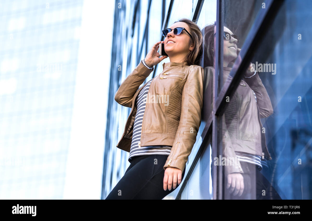 Woman talking on the phone  in city and leaning against glass business office building. Happy lady having conversation. Stylish person. Stock Photo
