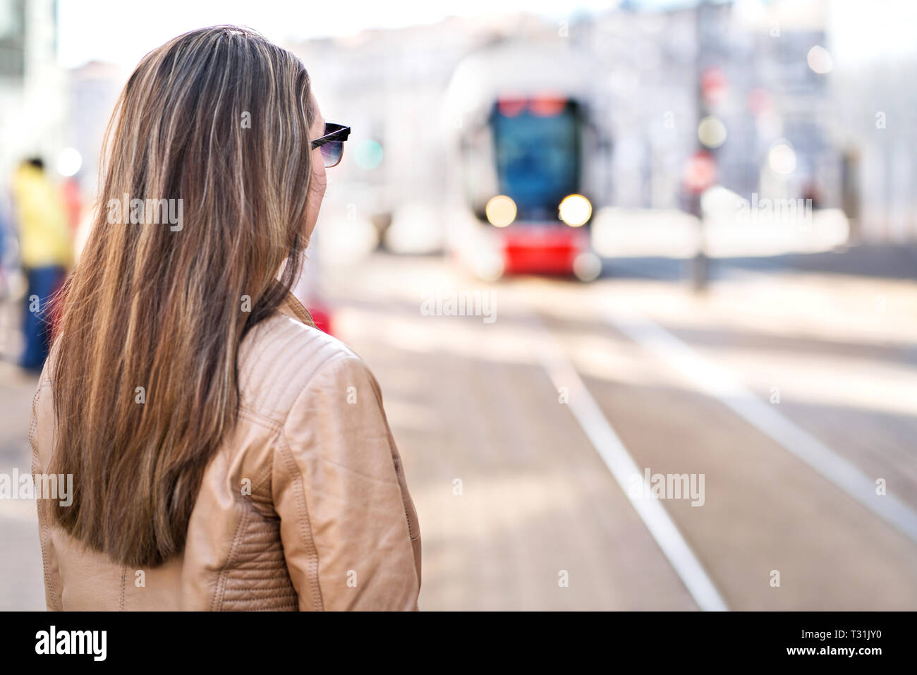 Back view of woman waiting for tram in stop. Passenger looking at arriving city train at station. Commuter or student or using public transportation. Stock Photo