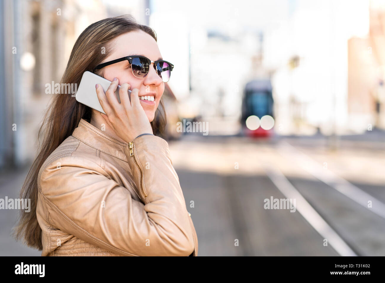 Young woman waiting for city train and talking on the phone. Passenger at tram stop calling with smartphone. Public transport and communication. Stock Photo