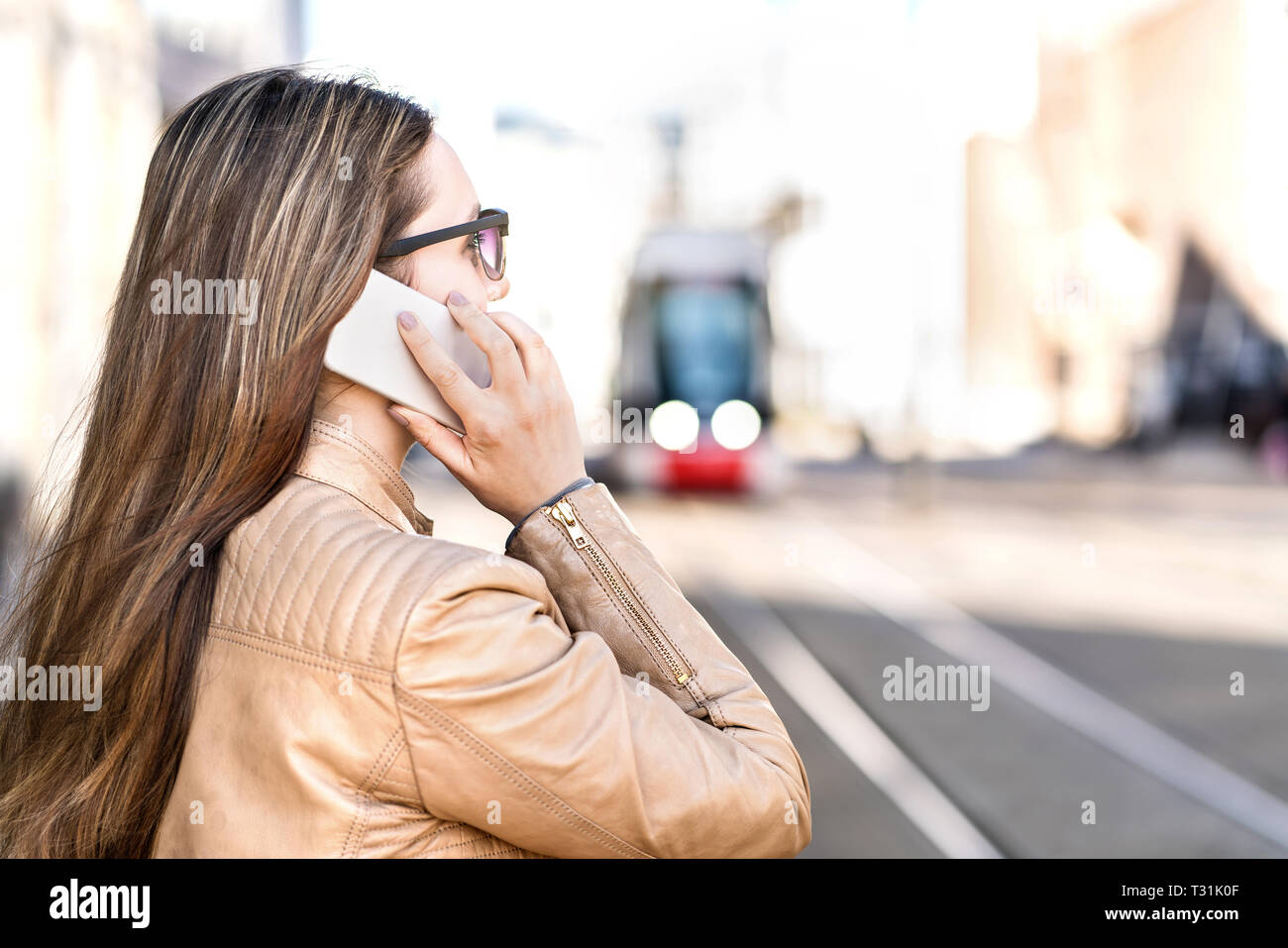 Busy businesswoman having a phone call while waiting for tram. Young woman with smartphone standing in city street. Electric train in the background. Stock Photo