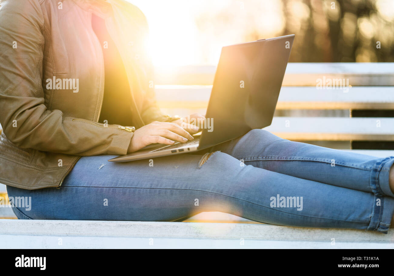Woman using laptop in sunset outdoors. Lady writing with computer while sitting on park bench in sunset. Remote work or studying in nature concept. Stock Photo