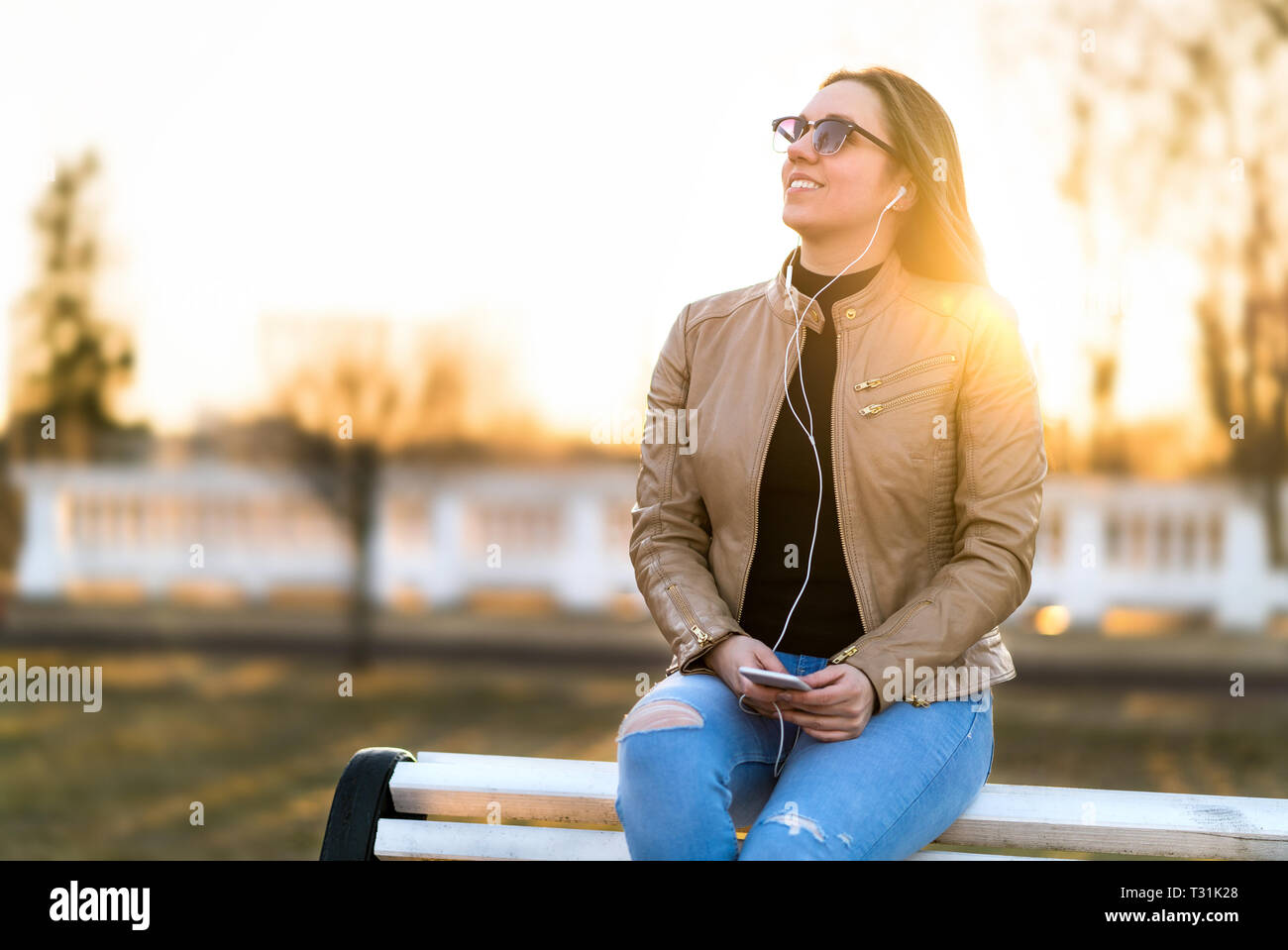 Woman listening to music outdoors. Happy smiling lady sitting on park bench holding mobile phone in sunset. Listening to audiobook outside. Stock Photo