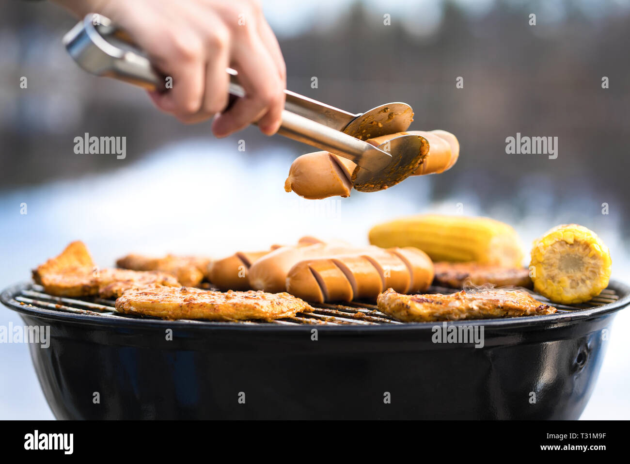 Person grilling with charcoal kettle grill full of delicious food. Cooking in outdoor kitchen in nature. Man or woman holding sausage. Stock Photo