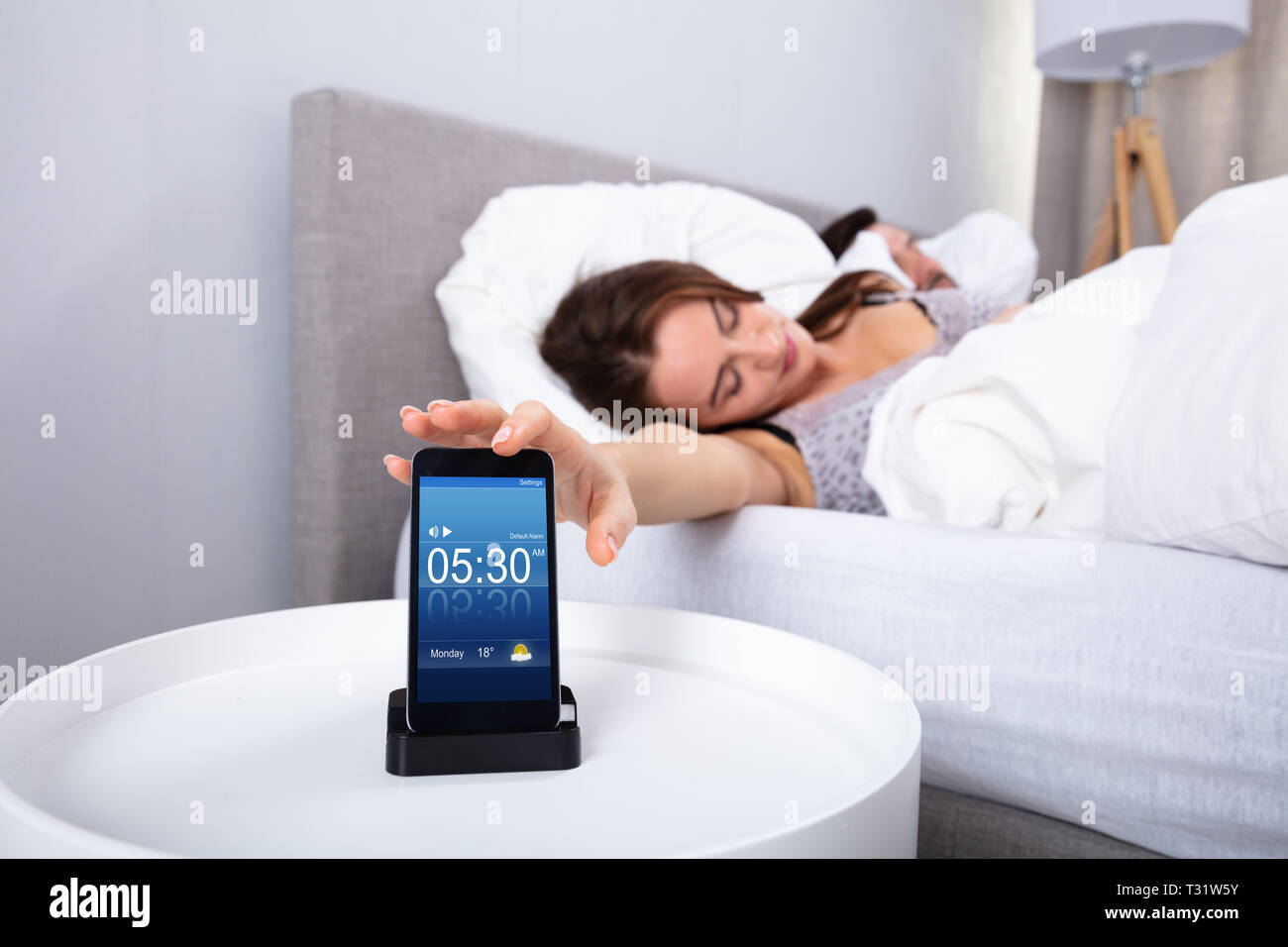 Young Woman Sleeping Near Alarm Set On Mobile Phone In Bedroom Stock Photo