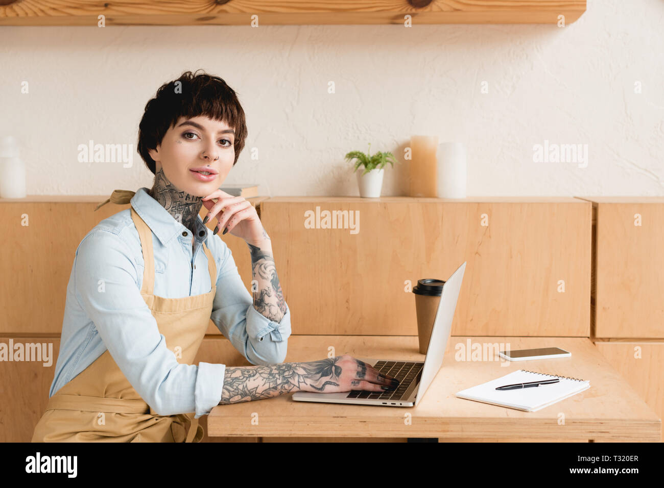 smiling waitress using laptop while sitting at table and looking at camera Stock Photo