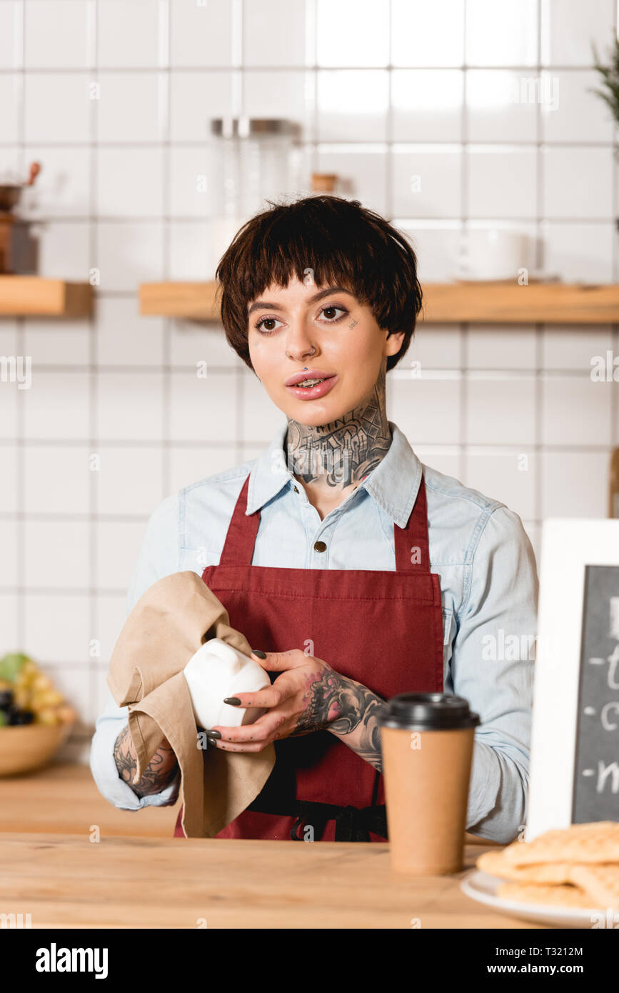 attractive barista in apron wiping cup while standing near bar counter