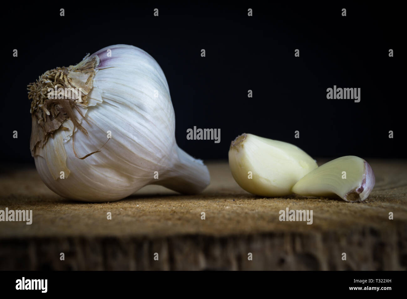 Two Garlic Cloves and Garlic Bulb on a wooden plate and dark background ...