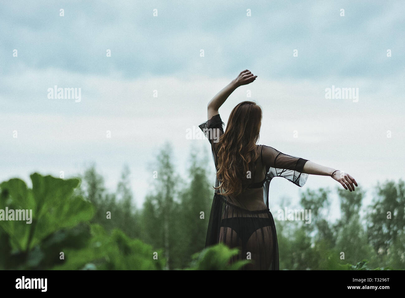 A dark-haired woman in a translucent black dress dancing against the sky and green grass. Relaxation and dreams. Stock Photo