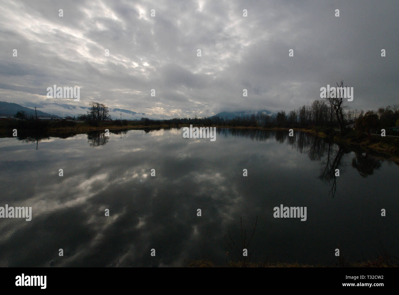 A placid backwater of the Vedder River reflects a turbulent gray overcast sky. Stock Photo