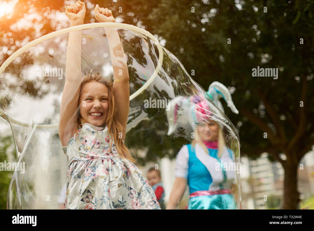 Lovely girl holding hands up and smiling while standing inside a large bubble during the festival on a sunny day in the park Stock Photo