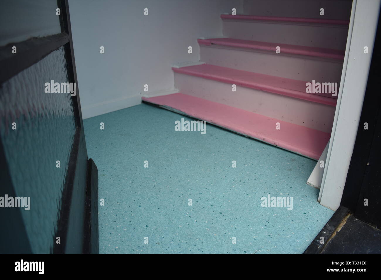 A glass door opens to a lino floor and pink painted wooden stairs in an older apartment building Stock Photo