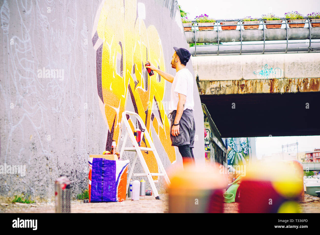 Street artist painting a colorful graffiti on a grey wall under bridge - Young graffiter writing and drawing murales with yellow spray Stock Photo