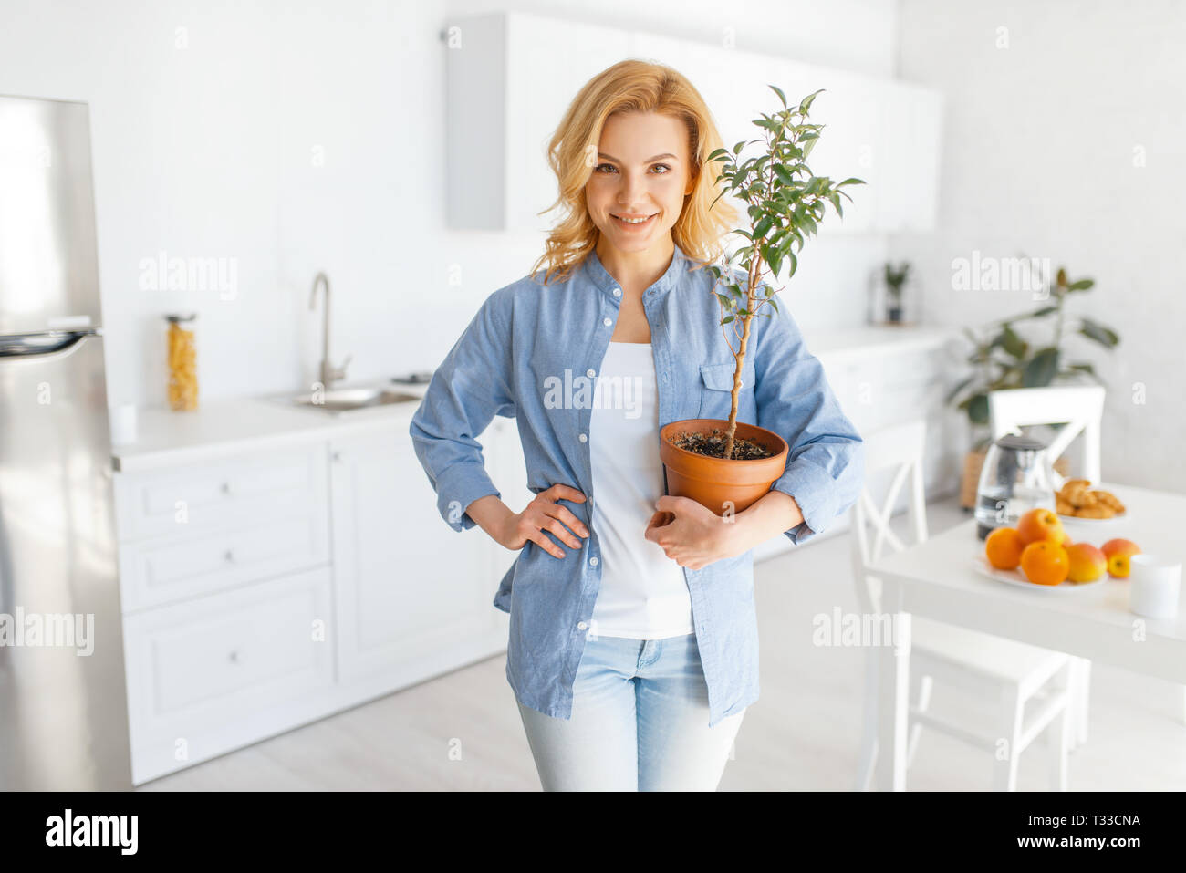 Young woman holds a flower in a pot on the kitchen with snow-white interior. Female person preparing breakfast at home in the morning, healthy nutriti Stock Photo