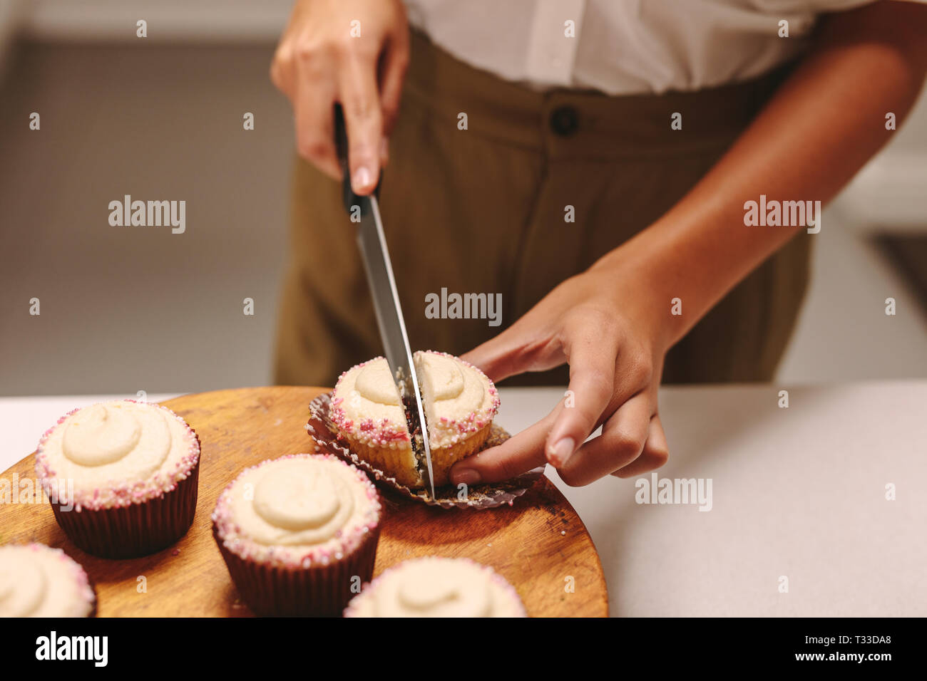 Close up of pastry chef cutting a homemade cupcake on wooden board with knife. Woman confectioner preparing muffins in the kitchen. Stock Photo