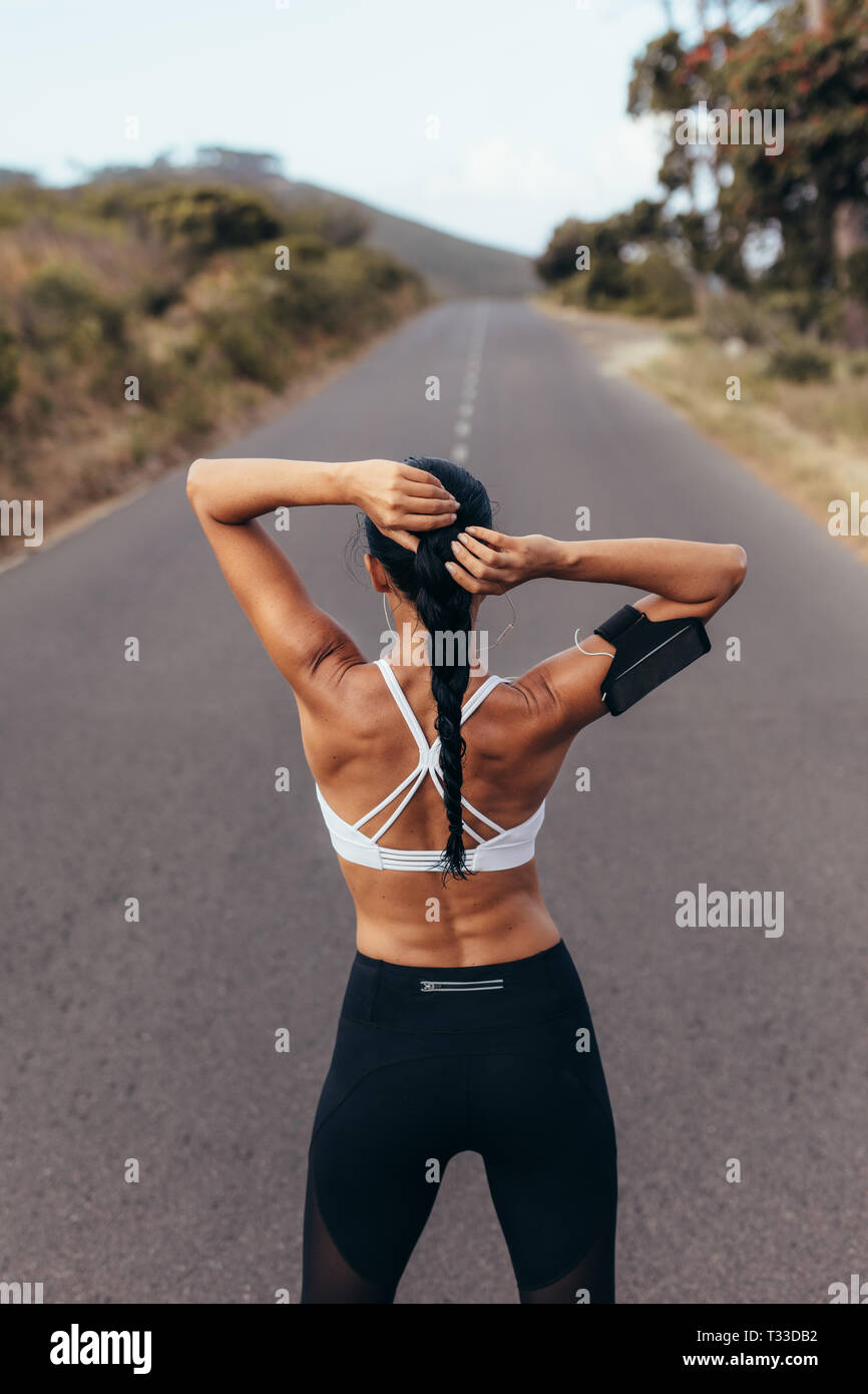 Rear view shot of fit young woman tying her hair and getting ready for a running workout. Female athlete before a run on empty road. Stock Photo