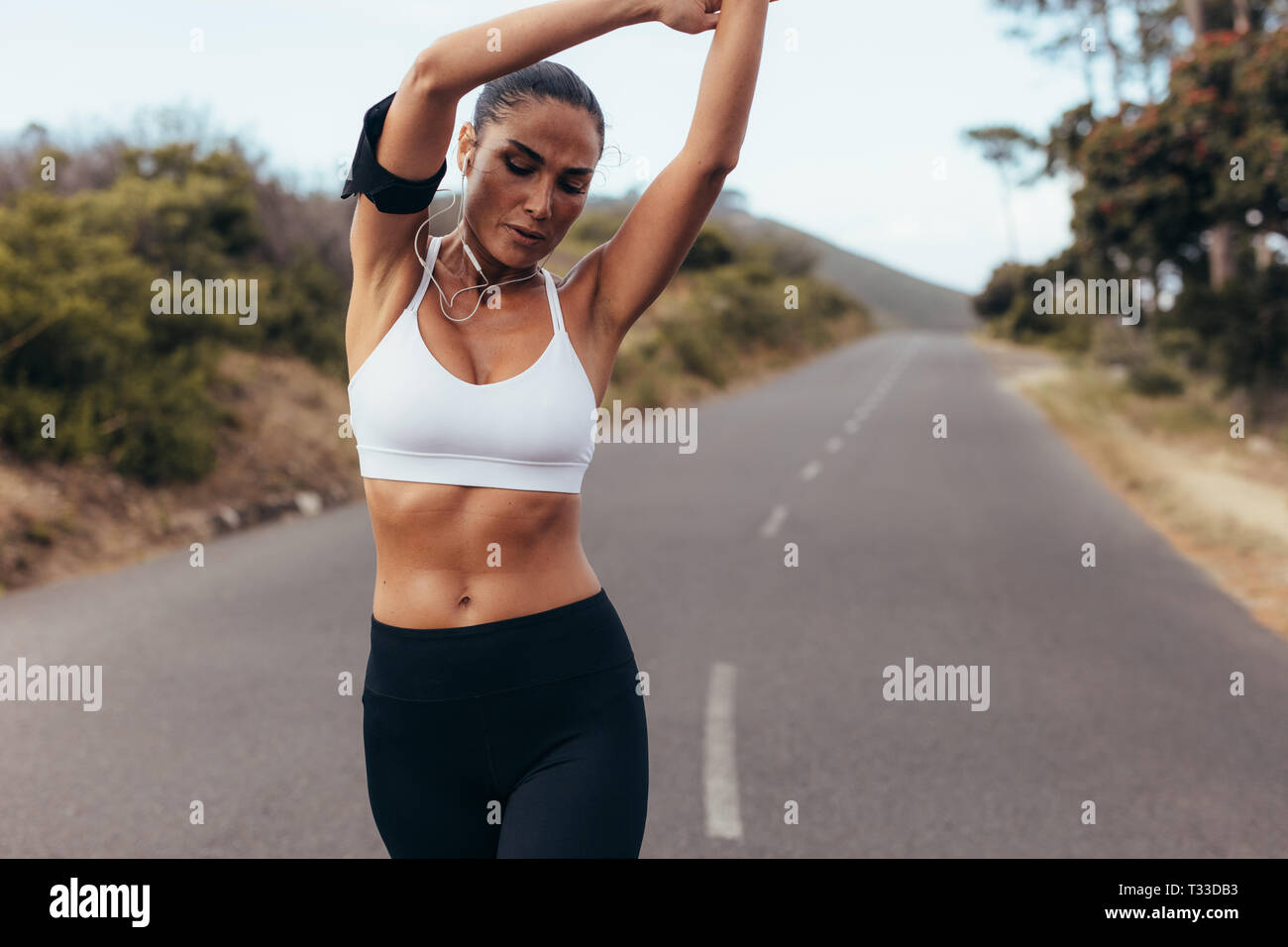 Sporty woman stretching her arms while walking on a empty road in morning. Female runner relaxing her muscles while working out outdoors. Stock Photo