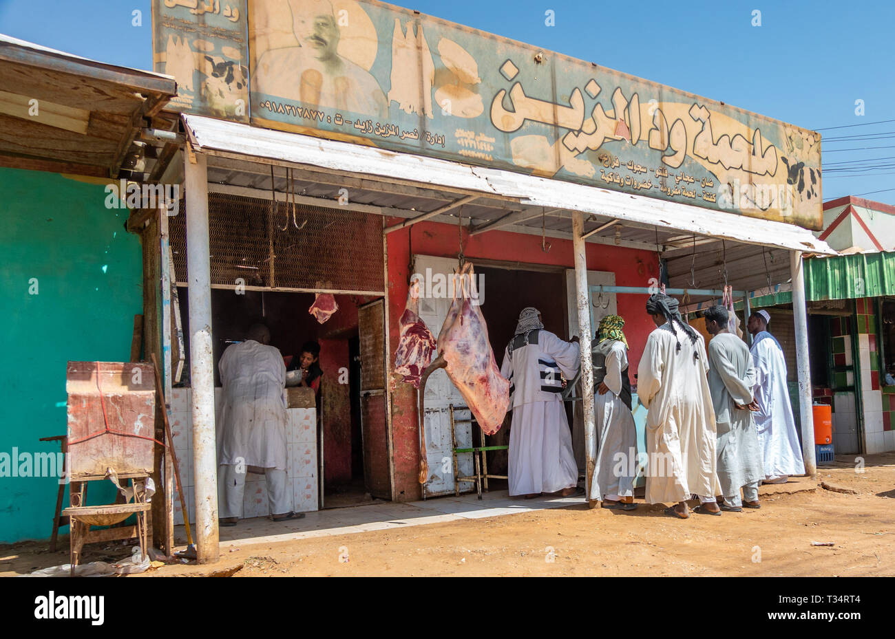 Nuri, Sudan, February 9., 2019: A large piece of beef hangs in front of a butcher's while men negotiate with the butcher in caftans and turban. Stock Photo