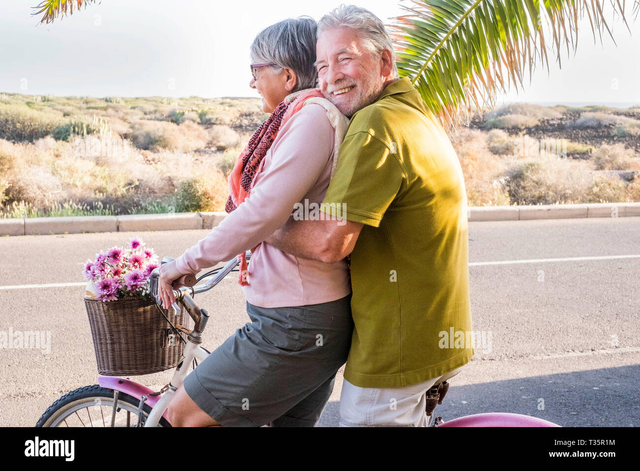 beautiful aged couple of senior in vacation go both on an old bike and enjoy the summer sunlight. tropical place and country side desert in background Stock Photo