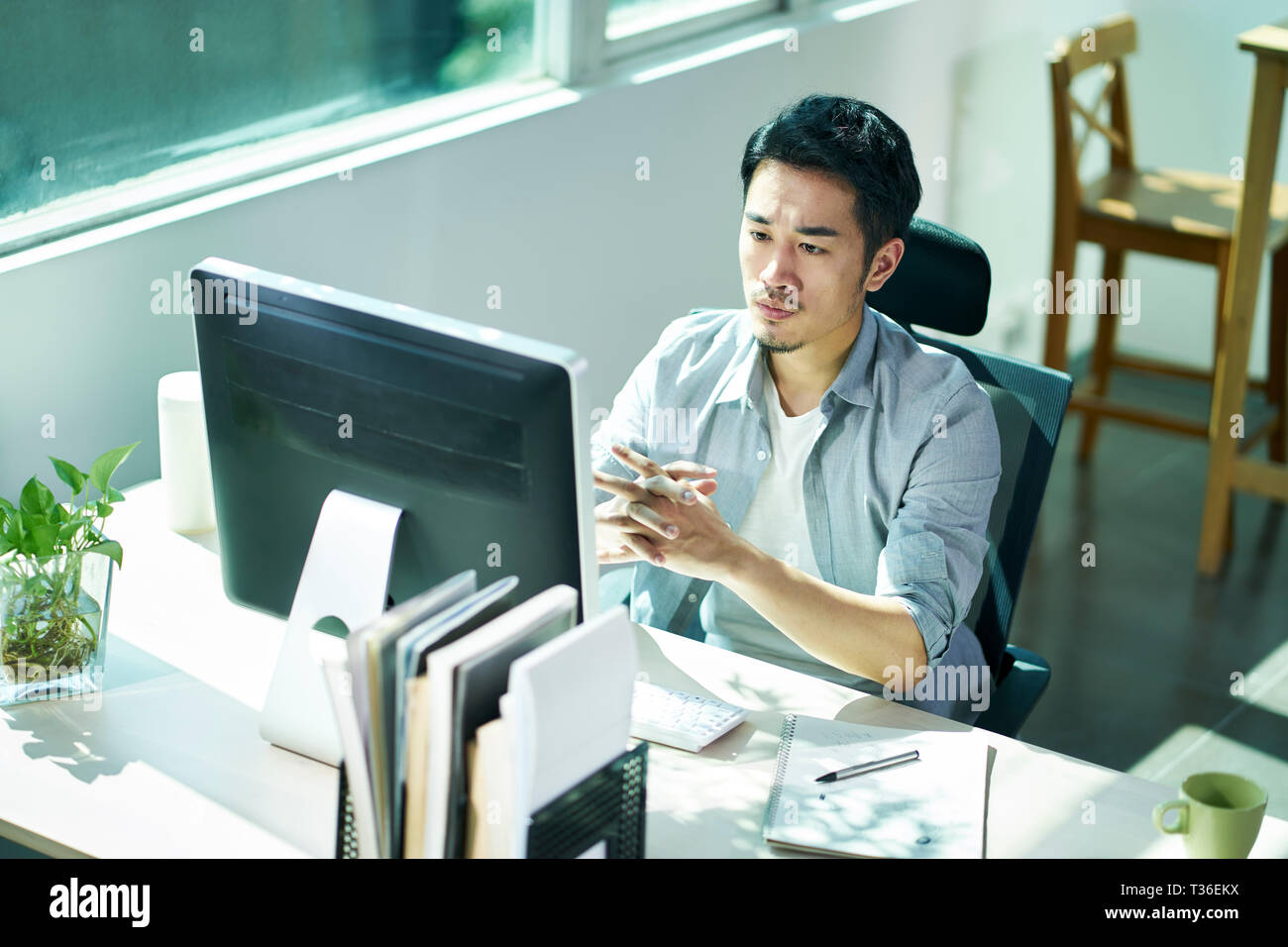 young asian business person sitting in office thinking while looking at monitor of desktop computer. Stock Photo