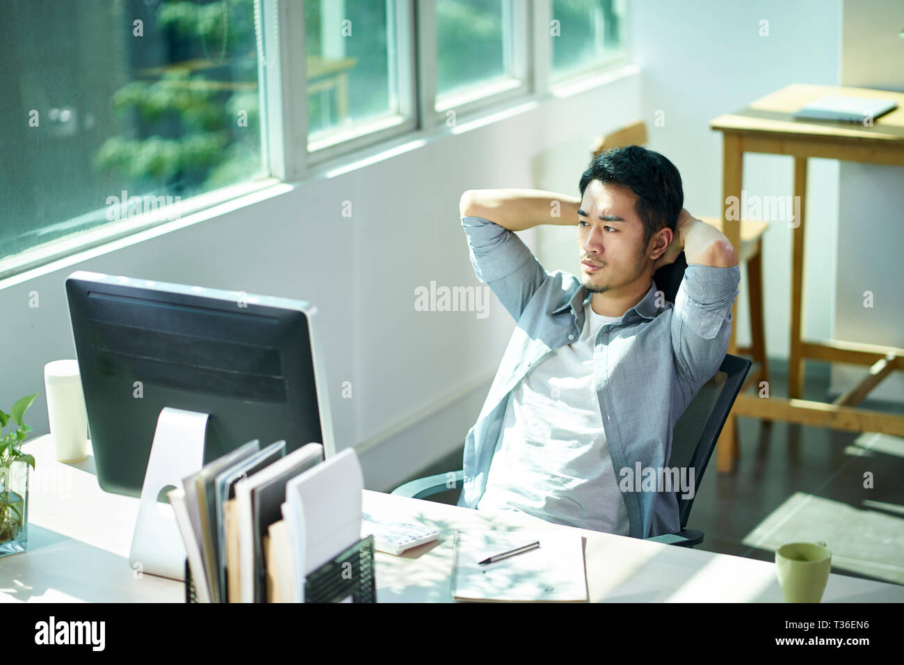 young asian business man entrepreneur contemplating in office, hands behind head frowning. Stock Photo