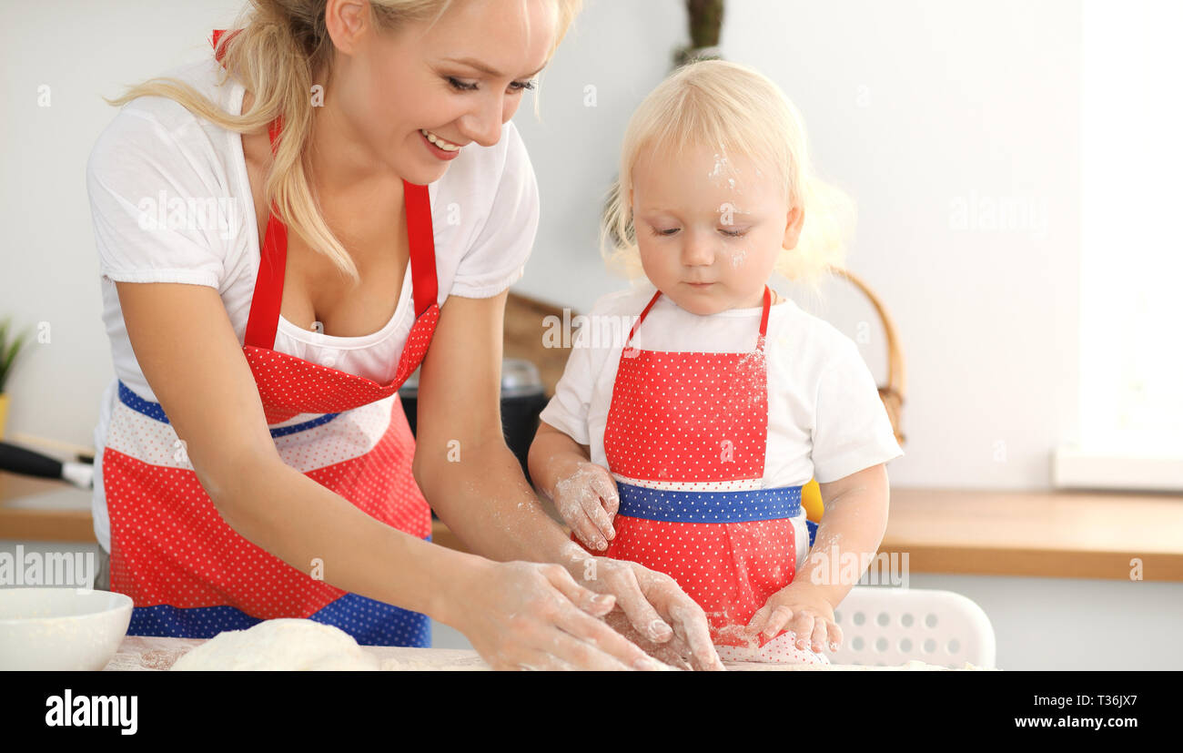 Little girl and her blonde mom in red aprons playing and laughing while kneading the dough in kitchen. Homemade pastry for bread, pizza or bake Stock Photo