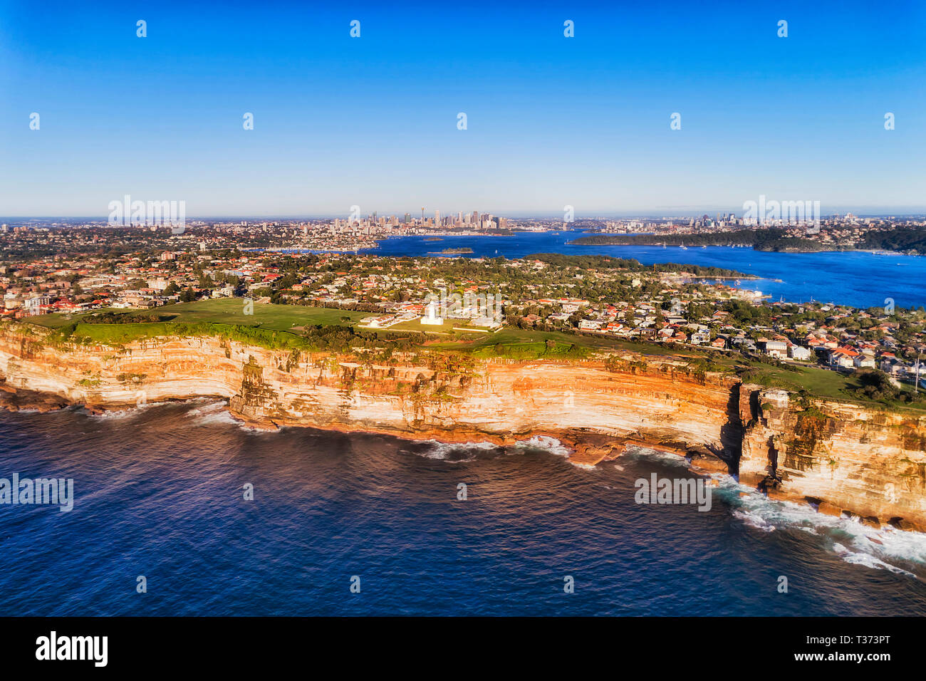 Sandstone cliff edge of Sydney Eastern suburbs facing Pacific ocean in aerial view over distant Harbour and city CBD on horizon. Stock Photo