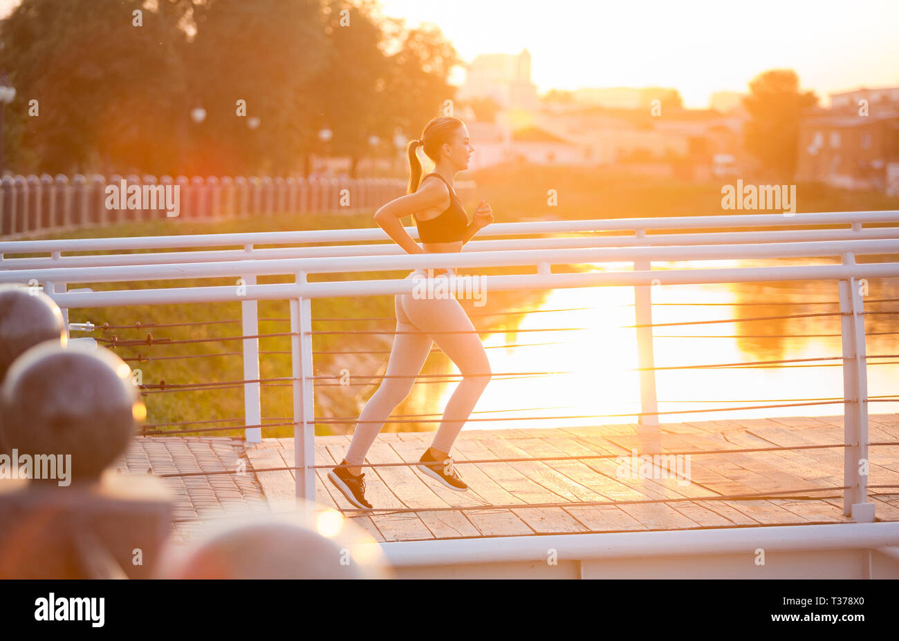 Active lifestyle. Millennial fitness girl jogging on bridge early in morning, sun flare, copy space Stock Photo