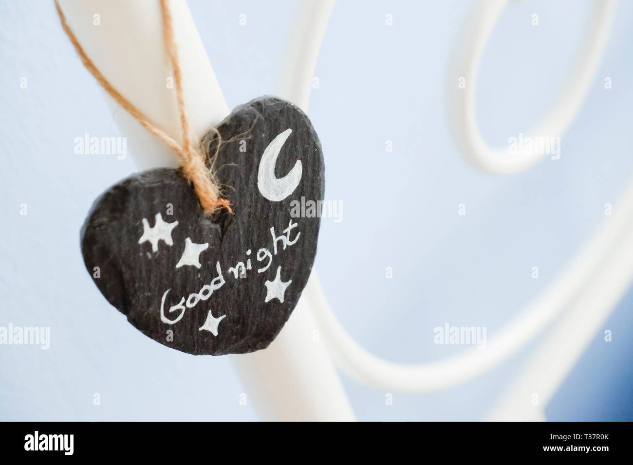 small heart of slate hangs with a rope  as decoration on a steel frame bed with goognight written on it and a drawn moon with stars Stock Photo