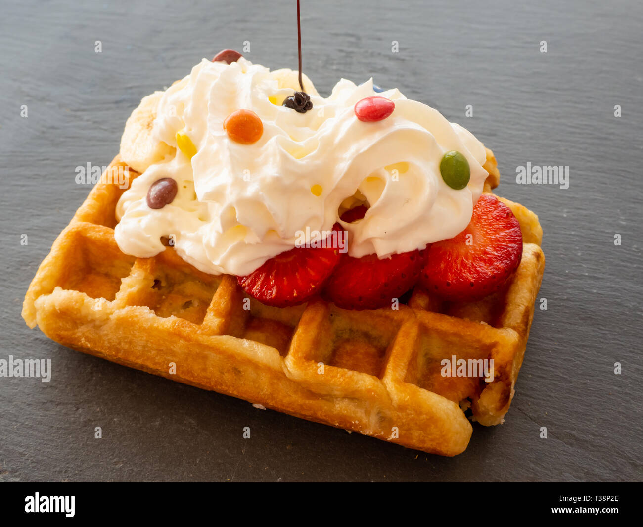 Chocolate syrup falling on a Belgian waffle with cream and colorful chocolates on pieces of strawberry on a slate plate Stock Photo