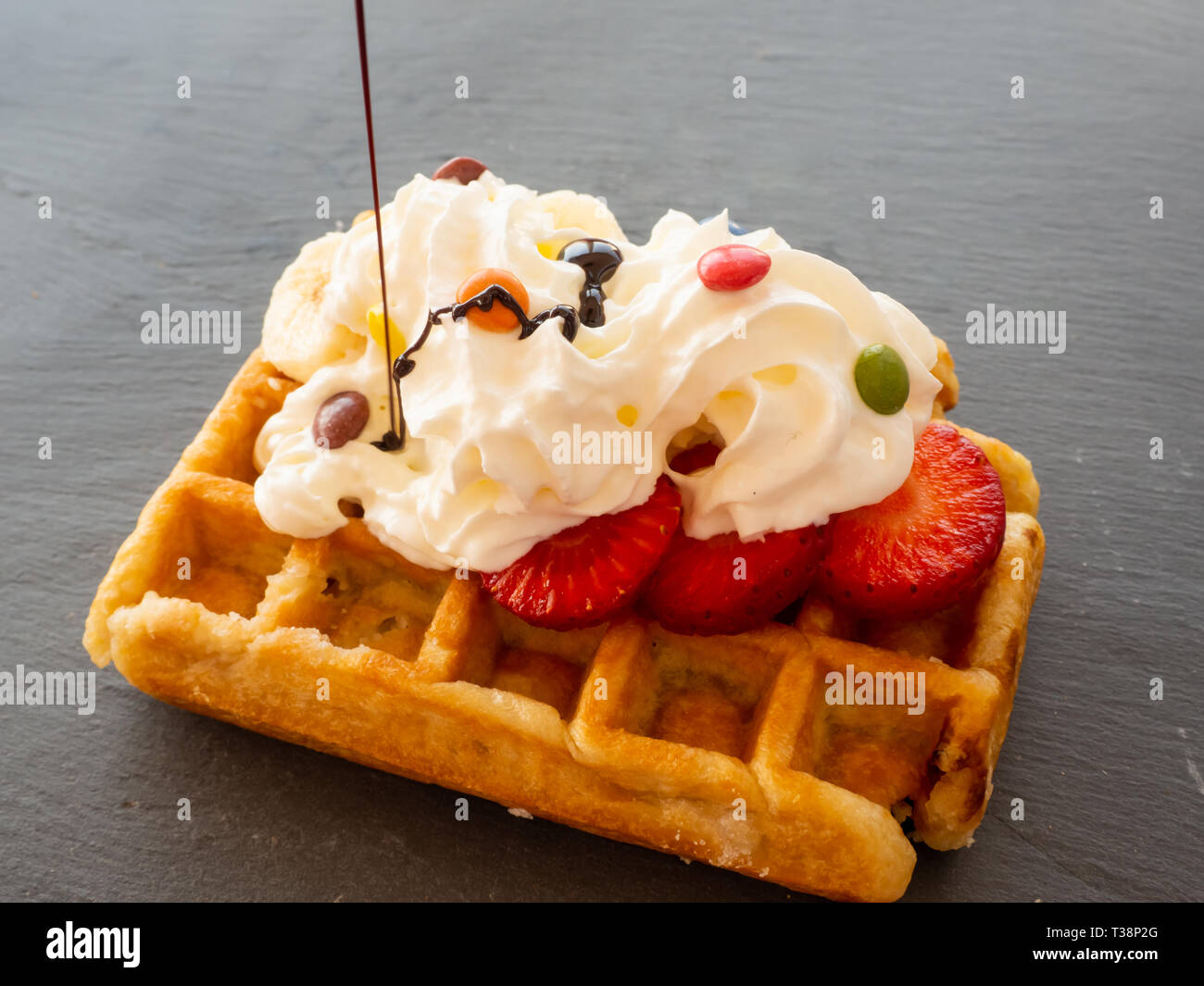 Chocolate syrup falling on a Belgian waffle with cream and colorful chocolates on pieces of strawberry on a slate plate Stock Photo