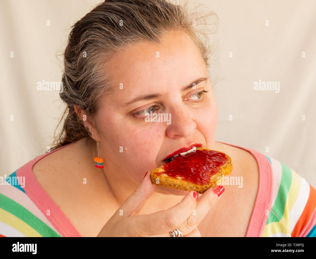 An adult woman with red nails and lips eating a rusk with raspberry jam Stock Photo