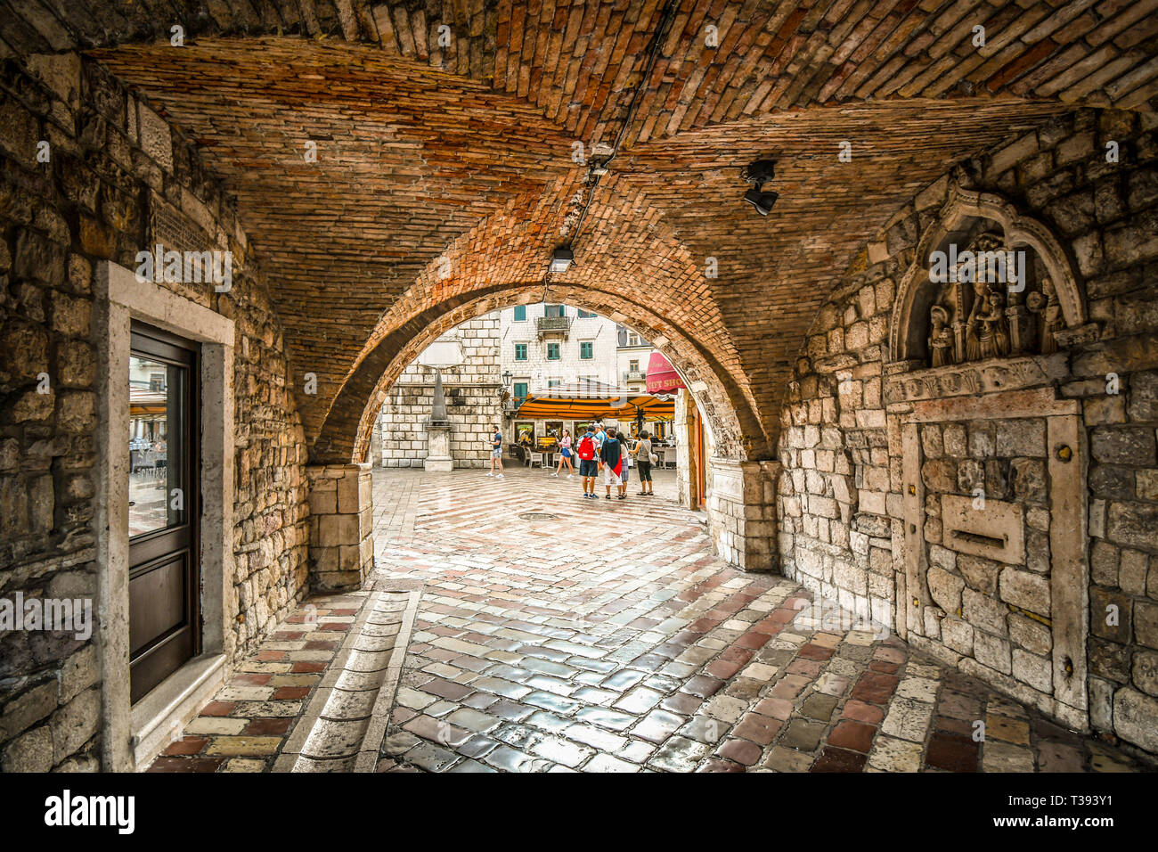 The Square of the Arms opens out in front as tourists enter the gated, walled medieval fortress town of Kotor, Montenegro through it's tunnel entrance Stock Photo