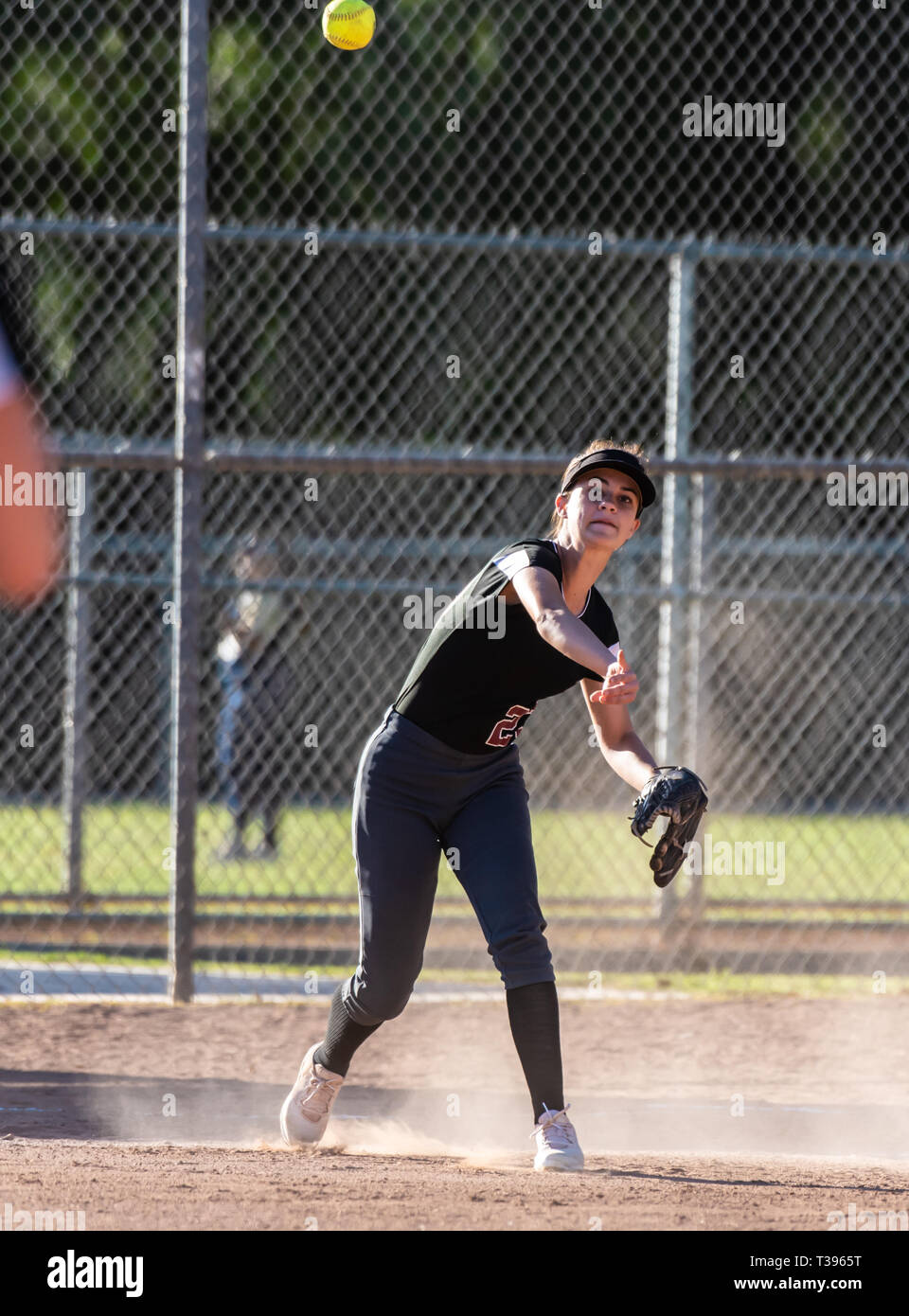 Female teenage softball player in black uniform throwing ball across infield for the out in a cloud of dust. Stock Photo