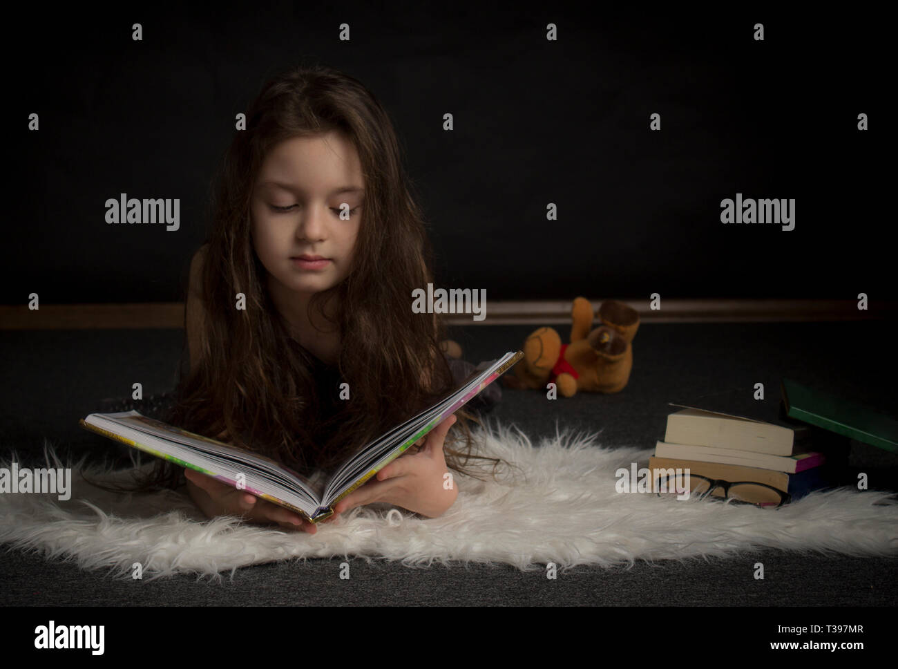 Longhair little girl reading a book in a dark room. Stock Photo