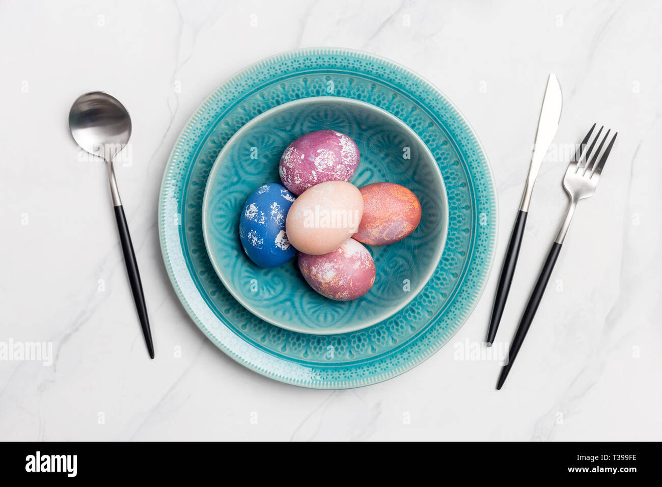 Table setting with colorful Easter eggs in blue plate on white marble background. Concept of Holiday dinner. Flat lay, table top Stock Photo